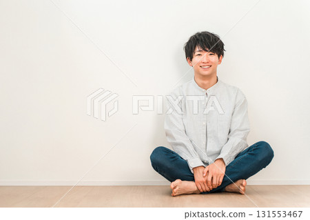 A young Asian man sitting cross-legged in a living room with white walls A young Asian man sitting cross-legged in a living room with white walls 131553467