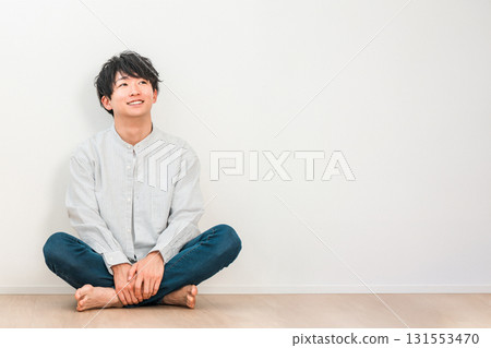 A young Asian man sitting cross-legged in a living room with white walls (Imagination, Dream, Thinking, Conception, Plan) 131553470