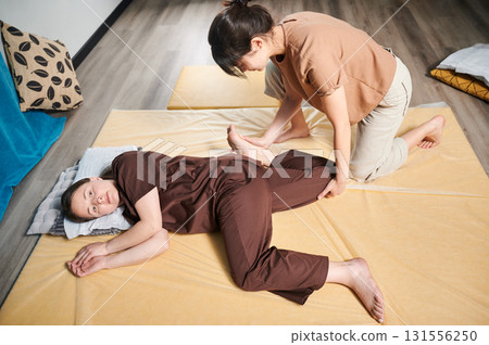 Masseuse making thai yoga massage. Woman therapist doing traditional massage treatment, stretching female patient's body. Client lying on mat, enjoying hand massage in calm atmosphere. 131556250