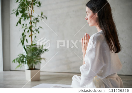 Beautiful woman in white dress sitting on mat in lotus position before Thai massage, practicing yoga, showing namaste sign. 131556256