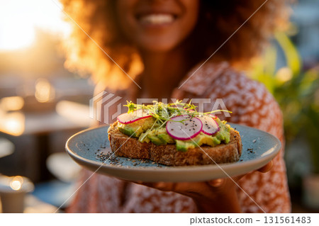 Close-up of a woman holding a sandwich with toasted bread sliced avocado green herbs and red radishes seasoned with pepper flakes. 131561483
