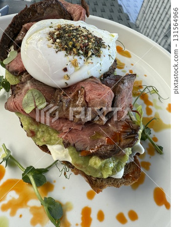 Gourmet sandwich with medium rare beef slices poached egg avocado cheese nuts and microgreens served on dark bread on white plate glass table outdoor daylight Gourmet sandwich with medium rare beef slices poached egg avocado cheese nuts and microgreens served on dark bread on white plate glass table outdoor daylight 131564966