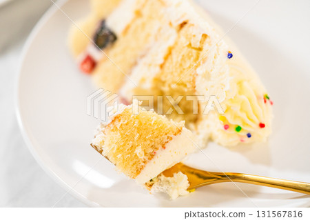 A detailed close-up of a golden fork lifting a bite-sized piece from a layered cake slice topped with creamy frosting and rainbow sprinkles. The soft cake texture and piped decoration are clearly A detailed close-up of a golden fork lifting a bite-sized piece from a layered cake slice topped with creamy frosting and rainbow sprinkles. The soft cake texture and piped decoration are clearly 131567816
