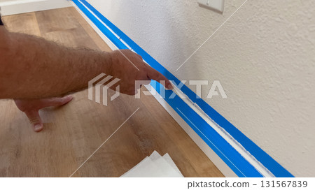 A close-up of a person's hand pointing to blue painter's tape applied to a wall baseboard, highlighting preparation work for painting the trim in a residential setting. 131567839
