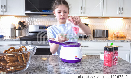 With focused attention, this budding culinary artist dips pretzels into a pot of melted chocolate, creating sweet delights in the warmth of a well-lit home kitchen. 131567841