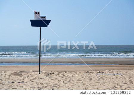 Wooden model of boat on pole on sandy beach with real big cargo ship in background, sunny day 131569832