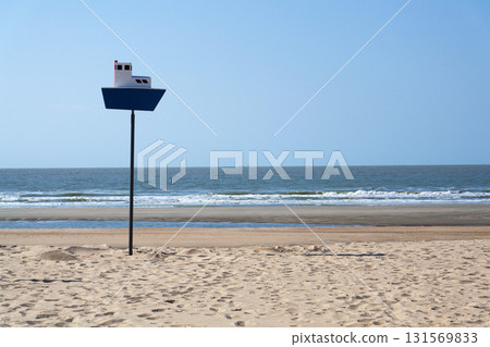Wooden model of boat on pole on sandy beach with real big cargo ship in background, sunny day 131569833