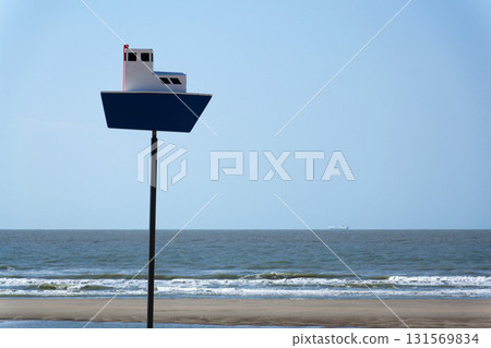 Wooden model of boat on pole on sandy beach with real big cargo ship in background, sunny day 131569834
