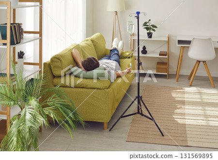 Young man receiving IV drip infusion and vitamin therapy lying on the couch at home. 131569895