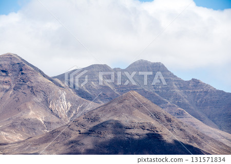 Large bare mountains with visible geological layers cast in the shadows of clouds on Fuerteventura in the Canary Islands Large bare mountains with visible geological layers cast in the shadows of clouds on Fuerteventura in the Canary Islands 131571834