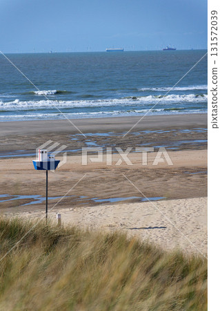 Wooden model of boat on pole on sandy beach with real big cargo ship in background, sunny day 131572039