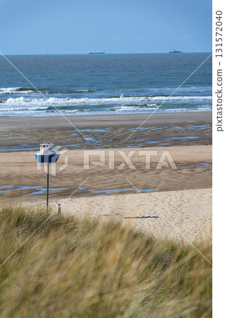 Wooden model of boat on pole on sandy beach with real big cargo ship in background, sunny day 131572040