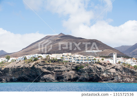 A white resort hotel standing on a cliff visible from the sea and a brown bald mountain with visible geological strata on Fuerteventura Island A white resort hotel standing on a cliff visible from the sea and a brown bald mountain with visible geological strata on Fuerteventura Island 131572536