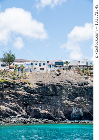 A white resort hotel standing on a cliff visible from the sea and a brown bald mountain with visible geological strata on Fuerteventura Island 131572545