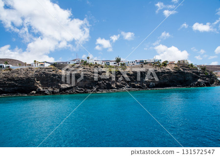 A white resort hotel standing on a cliff visible from the sea and a brown bald mountain with visible geological strata on Fuerteventura Island 131572547
