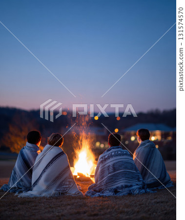 Group of four friends sit together around warm bonfire at night. People wrapped in blanket feel peaceful companionship during an outdoor gathering under twilight sky 131574570