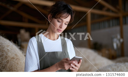 Focused woman farmer using smartphone inside rustic barn. small business owner manages inventory of wool with modern technology, looking concentrated and professional 131574600