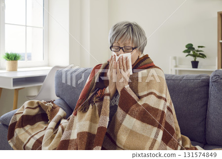 Sick upset senior woman with runny nose wrapped in a blanket sitting at home. Sick upset senior woman with runny nose wrapped in a blanket sitting at home. 131574849