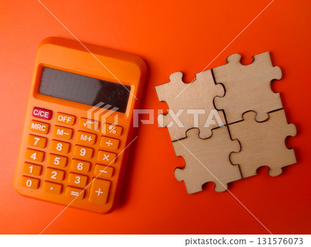 The image displays a small orange electronic calculator alongside four interlocking wooden puzzle pieces on an orange background. 131576073