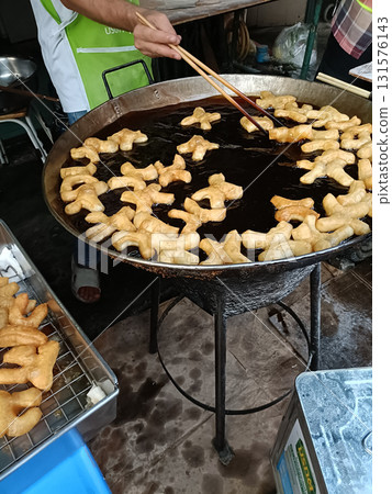 Freshly fried dough pastries in large round wok over hot oil, vendor using chopsticks to turn golden cruller pieces with busy market cooking atmosphere Freshly fried dough pastries in large round wok over hot oil, vendor using chopsticks to turn golden cruller pieces with busy market cooking atmosphere 131576143
