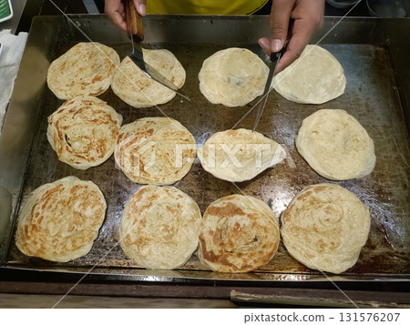 Flaky flatbread cooking on hot griddle with hands flipping pieces, warm golden layers and crisp texture inviting appetite 131576207