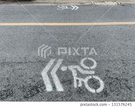 Bicycle lane symbol on asphalt road with chevron arrows, faded white marking and yellow centerline, urban street scene showing pavement texture and curb Bicycle lane symbol on asphalt road with chevron arrows, faded white marking and yellow centerline, urban street scene showing pavement texture and curb 131576245