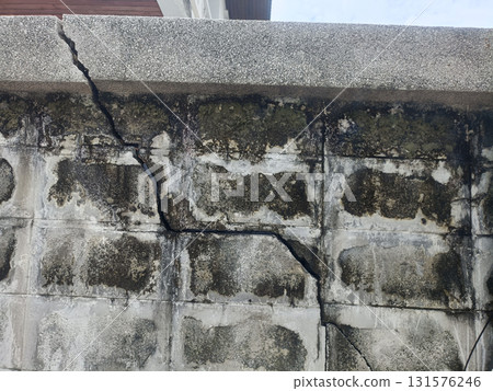 Cracked concrete wall with weathered stains and moss, vertical and diagonal fissure revealing structural damage and textured aged surface conveying decay and neglect Cracked concrete wall with weathered stains and moss, vertical and diagonal fissure revealing structural damage and textured aged surface conveying decay and neglect 131576246