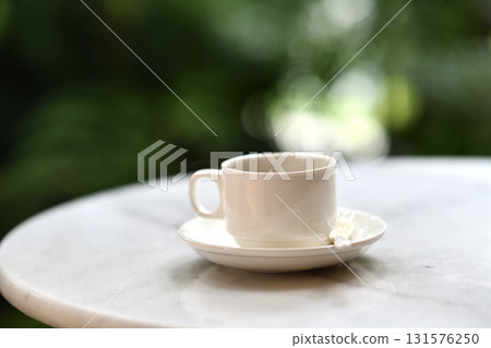 White ceramic cup on saucer with spoon on marble table in garden morning light, peaceful beverage moment for relaxing or breakfast 131576250
