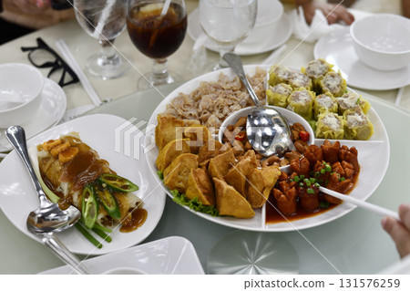 Steamed dumpling, fried wonton, sausage in sauce, rice roll with peanut sauce and sliced chili, assorted Chinese dim sum platter served on round table with glasses and bowls 131576259