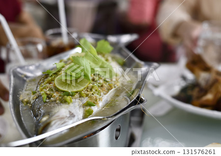 Steamed whole fish topped with lime slices, garlic, chili and fresh herb garnish served on metal platter, flavorful and aromatic presentation for dining enjoyment 131576261