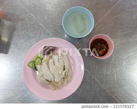 Boiled chicken served on seasoned rice with cucumber, blood pudding wedge and spicy dipping sauce beside clear soup, plated on pink dish and metal table conveying simple comforting street meal Boiled chicken served on seasoned rice with cucumber, blood pudding wedge and spicy dipping sauce beside clear soup, plated on pink dish and metal table conveying simple comforting street meal 131576327