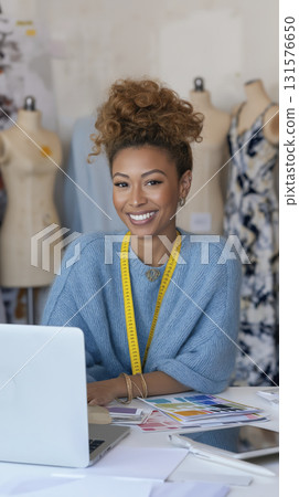 Smiling woman with curly hair wearing blue sweater and measuring tape around her neck, working fashion design studio filled with mannequins 131576650