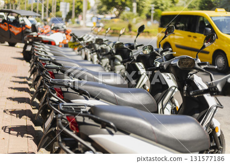 A row of parked scooters lines a busy city street. A yellow taxi waits nearby while sunlight shines on the scene. The atmosphere feels lively and urban A row of parked scooters lines a busy city street. A yellow taxi waits nearby while sunlight shines on the scene. The atmosphere feels lively and urban 131577186