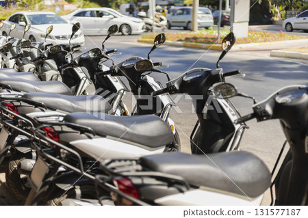 A row of scooters parked neatly along the roadside in a bustling city setting. Vehicles are visible in the background, showcasing the lively atmosphere of urban life A row of scooters parked neatly along the roadside in a bustling city setting. Vehicles are visible in the background, showcasing the lively atmosphere of urban life 131577187