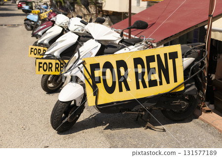 A row of scooters is parked outside a shop with large signs indicating they are available for rent. The sunny afternoon highlights the bustling atmosphere of the coastal area 131577189