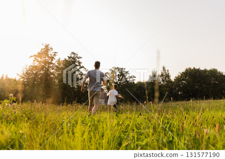 A father and his son run together through a lush green field during golden hour. Sunlight makes the scene warm and inviting as they share laughter and bonding moments 131577190