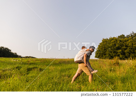 A father carries his son on his back as they explore a lush green field under a clear blue sky. They enjoy their quality time outdoors, connecting with nature and each other A father carries his son on his back as they explore a lush green field under a clear blue sky. They enjoy their quality time outdoors, connecting with nature and each other 131577222