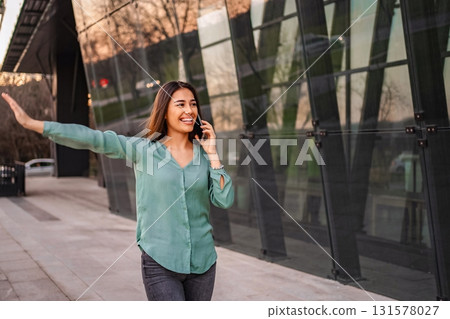 Young businesswoman waving while talking on the phone in front of a modern glass building 131578027