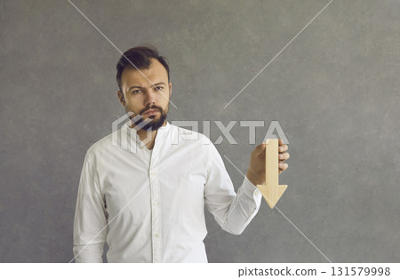 Sad business man with wooden down arrow in hand standing over studio background 131579998