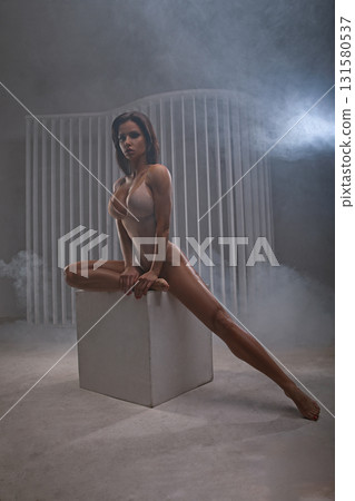 In a studio filled with soft smoke a beautiful woman strikes a graceful pose on one leg sitting on a cube. The lighting highlights her form and adds to the artistic atmosphere. 131580537