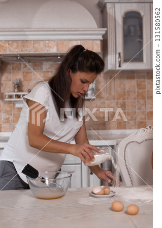 pregnant woman is whisking eggs with concentration. Flour dusts the countertop showing her involvement in cooking. The atmosphere feels inviting and nurturing. 131580562