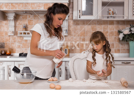 pregnant woman measures flour while her young daughter rolls dough. They share a fun moment creating a cake together with care and love. pregnant woman measures flour while her young daughter rolls dough. They share a fun moment creating a cake together with care and love. 131580567