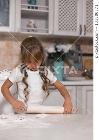 young girl rolls out dough with a wooden rolling pin. Flour covers the table as she focuses on making a delicious cake from scratch showcasing her baking skills. young girl rolls out dough with a wooden rolling pin. Flour covers the table as she focuses on making a delicious cake from scratch showcasing her baking skills. 131580571