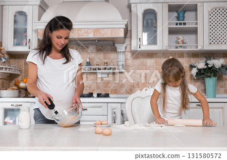 pregnant woman mixes batter in a bowl while her daughter rolls dough with a rolling pin. They are enjoying quality time together in their bright kitchen on a cheerful afternoon. pregnant woman mixes batter in a bowl while her daughter rolls dough with a rolling pin. They are enjoying quality time together in their bright kitchen on a cheerful afternoon. 131580572