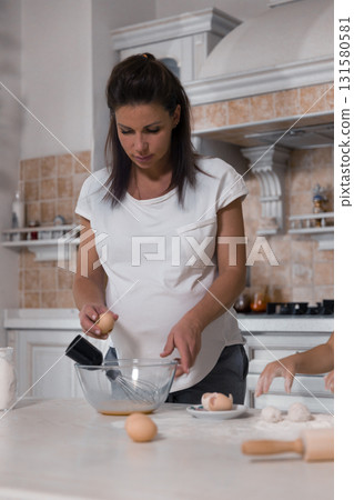 pregnant woman is mixing ingredients in a bowl. She carefully handles eggs as she prepares a meal surrounded by various cooking tools and a warm atmosphere. 131580581