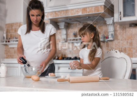 pregnant woman and her daughter mix ingredients to prepare a cake. The young girl rolls dough while her mother uses a mixer creating memories together. 131580582