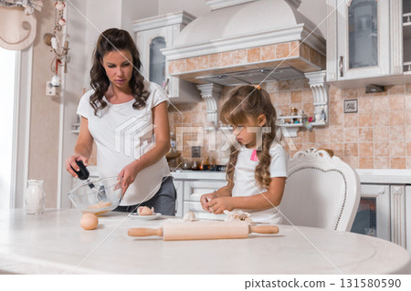 pregnant woman is mixing ingredients in a bowl while her little daughter is carefully rolling dough. They are both smiling and enjoying the moment. pregnant woman is mixing ingredients in a bowl while her little daughter is carefully rolling dough. They are both smiling and enjoying the moment. 131580590