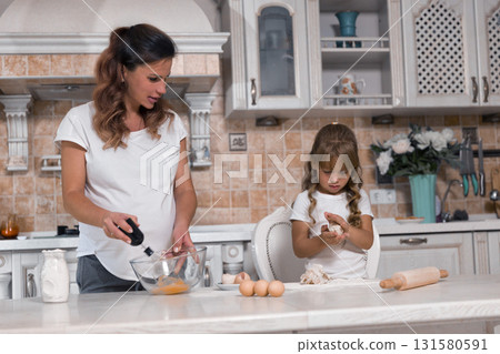 pregnant woman mixes ingredients in a bowl while her little daughter focused on her task kneads dough. Together they create sweet memories and a delicious cake. pregnant woman mixes ingredients in a bowl while her little daughter focused on her task kneads dough. Together they create sweet memories and a delicious cake. 131580591
