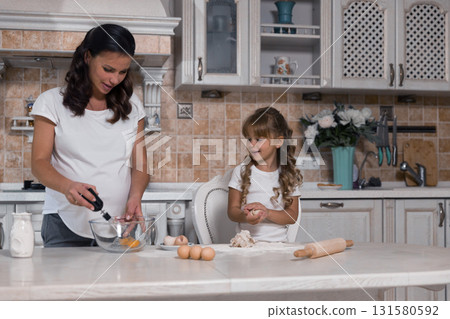 pregnant woman mixes ingredients in a bowl while her little daughter plays with dough. They share a joyful moment making a cake together. pregnant woman mixes ingredients in a bowl while her little daughter plays with dough. They share a joyful moment making a cake together. 131580592