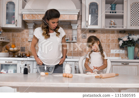 In a bright kitchen a pregnant woman and her young daughter are working together to bake a cake. They are mixing ingredients and creating memories as they enjoy their time together. 131580595
